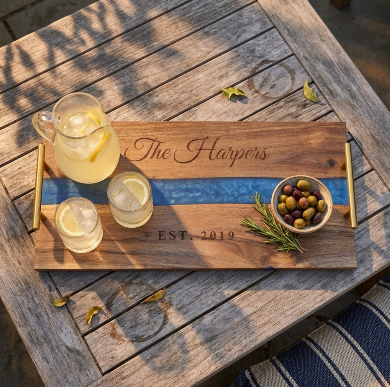 Blue river serving tray engraved The Harpers and EST. 2019 with a pitcher of lemonade, two glasses, olives and rosemary on a weathered teak patio table
