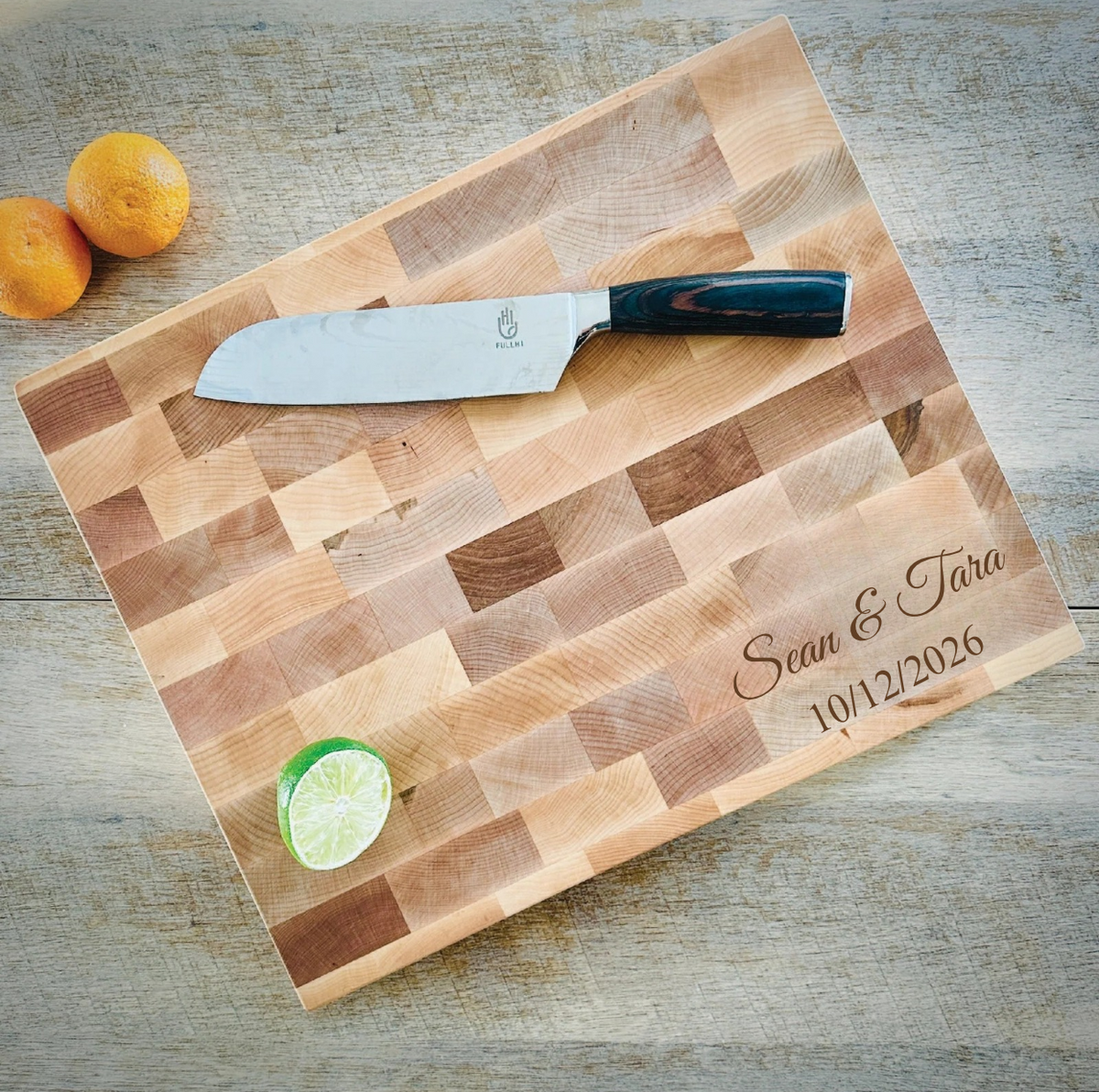 Maple Custom Butcher Block engraved Sean &amp; Tara in script with 10/12/2026 below, with a chef's knife, two oranges and a lime half on a gray wood table

