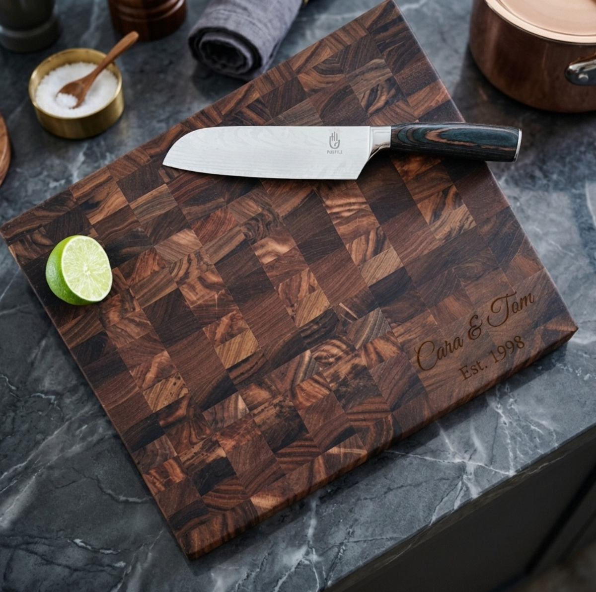 Walnut Custom Butcher Block engraved Cara &amp; Tom in script with Est. 1998 below, with a chef's knife and a lime half on a dark marble counter