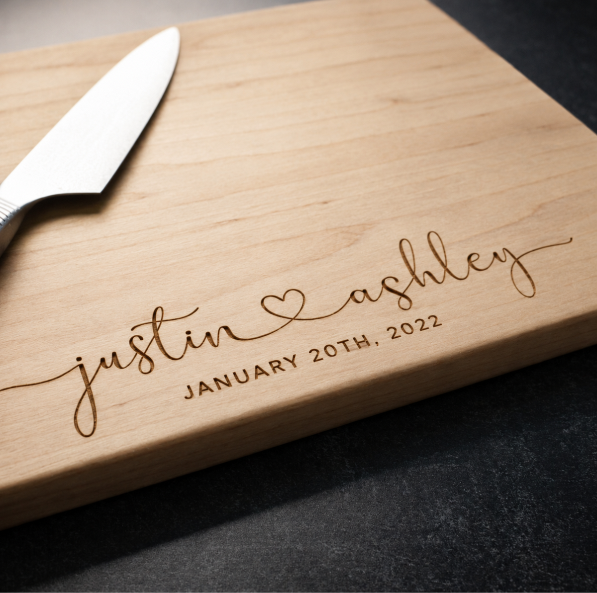 Close view of a maple cutting board engraved with justin and ashley in lowercase script joined by a heart and JANUARY 20TH, 2022, beside a chef's knife
