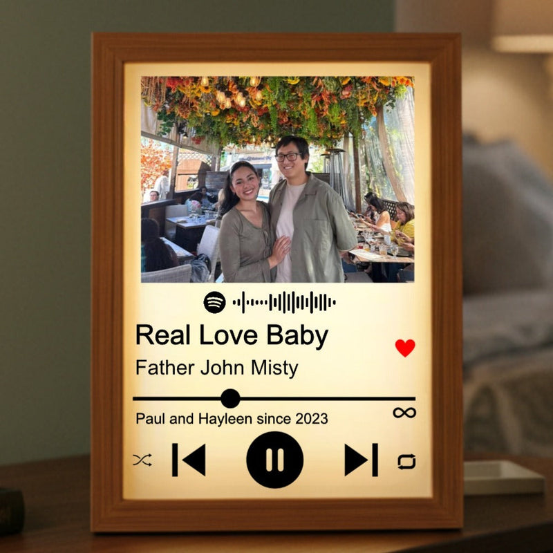 Wooden frame with warm backlight displaying couple photo, sample song "Real Love Baby" by Father John Misty, and personalized text "Paul and Hayleen since 2023" with Spotify player controls
