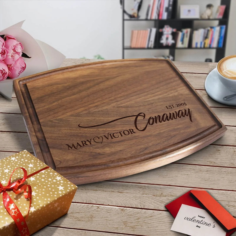Walnut cutting board engraved with sample names "Mary" and "Victor" and "Est. 2005," displayed on wooden table with coffee cup and pink flowers