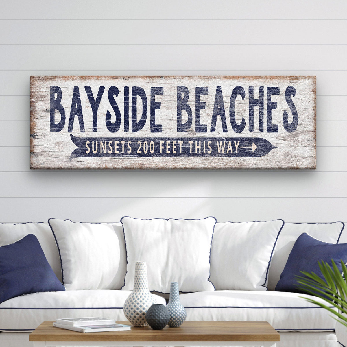 Saltwater Arrow Canvas displaying "Bayside Beaches" in bold navy distressed lettering above a navy directional arrow reading "Sunsets 200 Feet This Way," mounted above a white and navy sofa on a shiplap wall in a coastal living room | Willow &amp; Hive