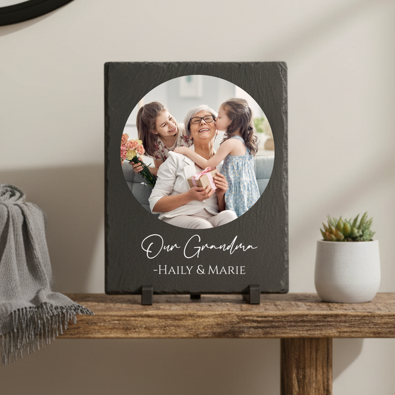 Dark slate plaque with circular photo cutout displaying a grandmother with two young girls holding flowers, personalized with sample text "Our Grandma" and names "Haily & Marie" below the image