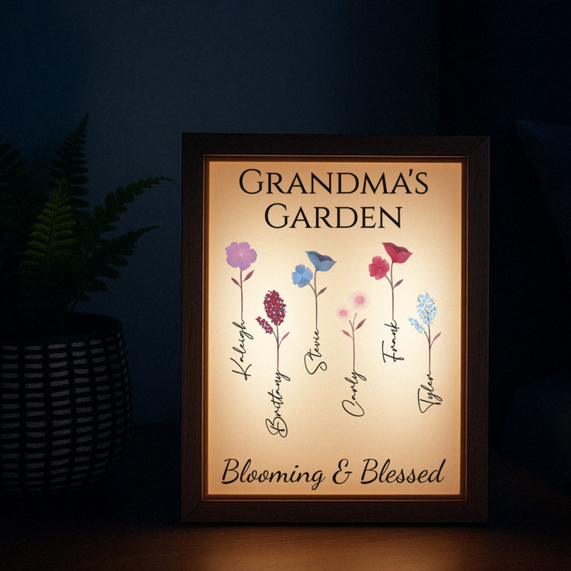 Backlit wood-framed garden sign displaying "Grandma's Garden" at top with five illustrated flowers labeled with sample names "Kathy," "Butterfly," "Steve," "Carl," and "Violet Freach," and "Blooming & Blessed" at bottom, glowing warmly against dark background.