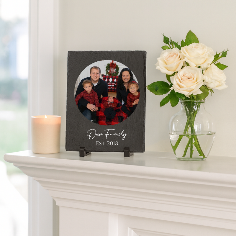 Dark slate frame with circular photo opening displaying family portrait, engraved with sample text "Our Family" and "Est. 2018" below, displayed on white mantel shelf next to candle and fresh roses