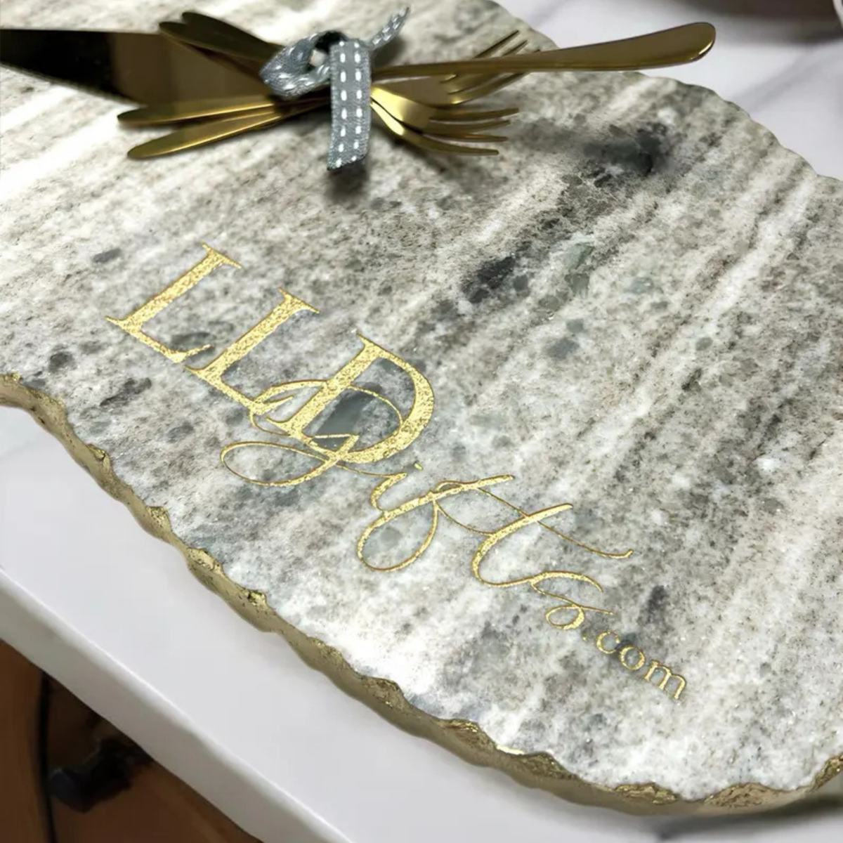 Marble charcuterie board engraved with sample text "LEXI" and "Est. 2024.com" in gold lettering, with brass serving utensils tied with gray ribbon on top