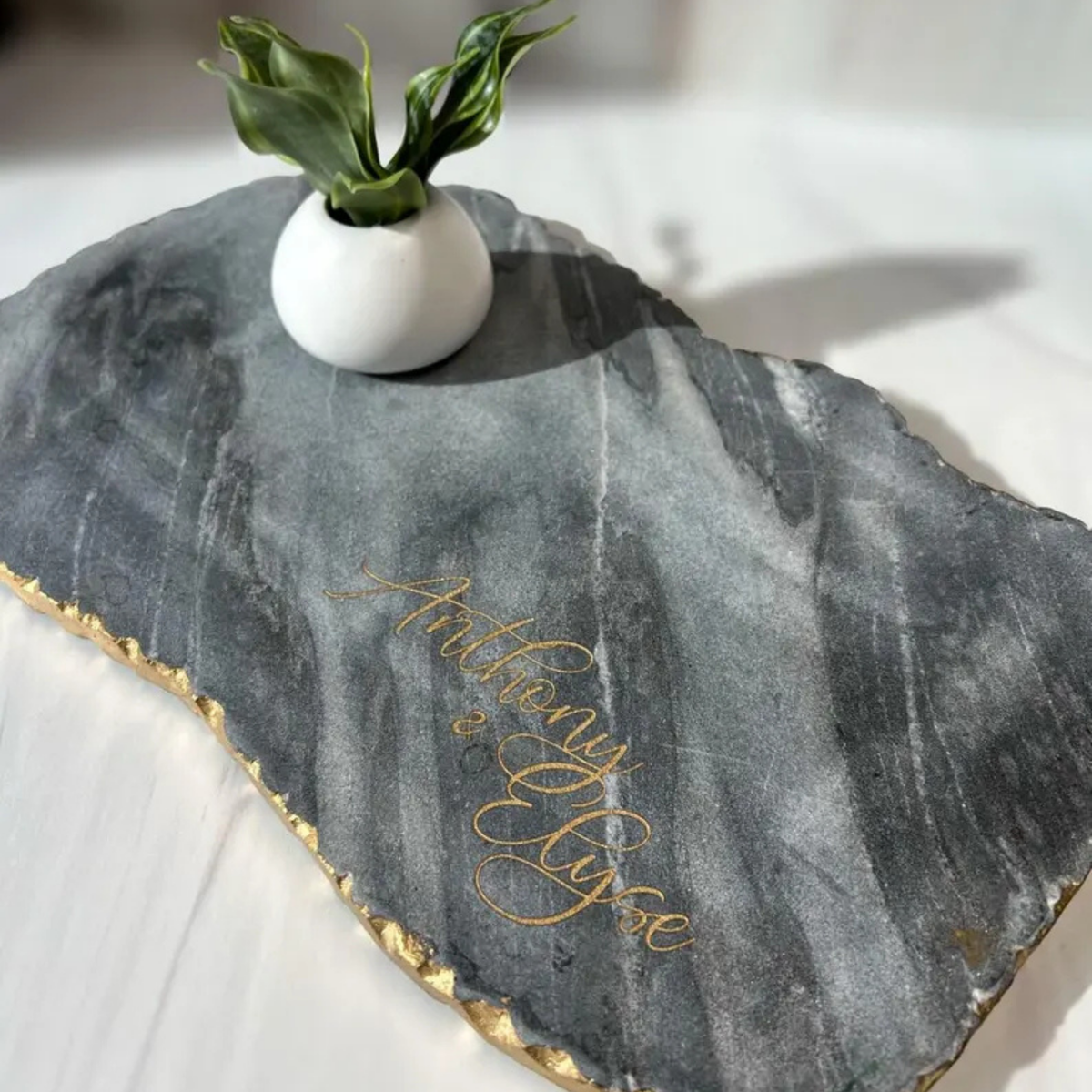 Dark gray marble board with gold-leaf edge, personalized with sample script engraving reading "Andrew &amp; Nicole," styled with small white vase holding green plant leaves.