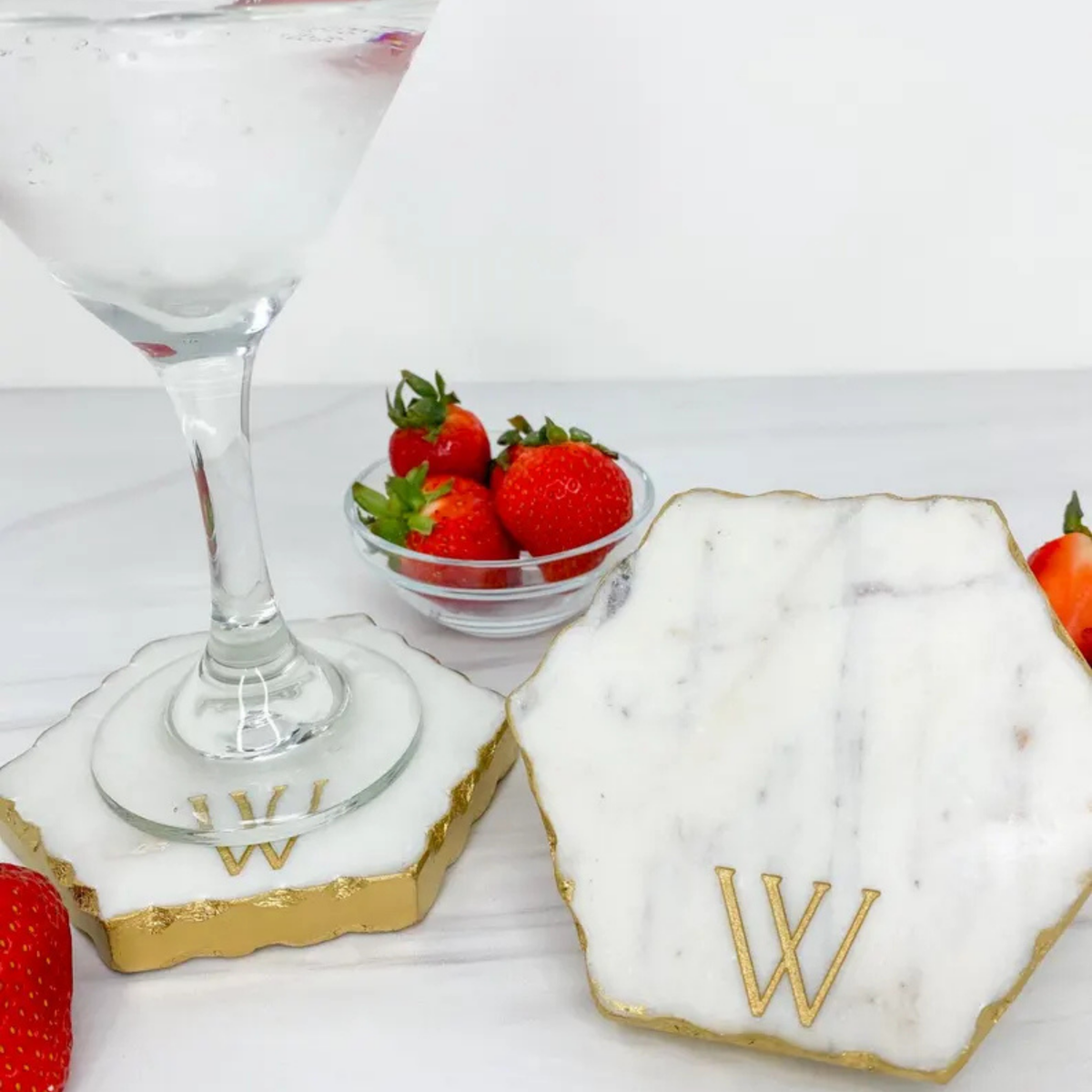 Marble charcuterie board with gold edge engraved with sample initial "W" positioned next to a cocktail glass and fresh strawberries on white surface