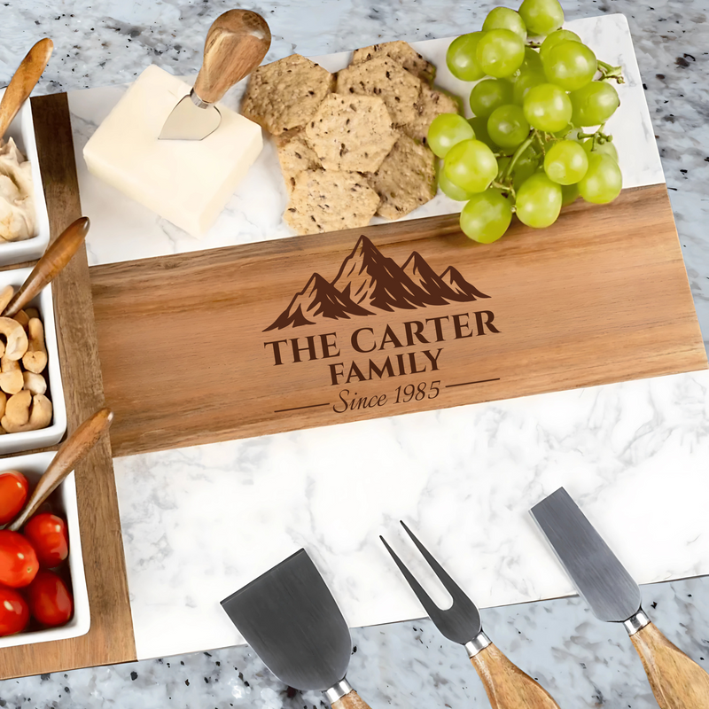 Wooden charcuterie board engraved with sample family name "THE CARTER FAMILY" and established year "Since 1985," styled with cheese, crackers, grapes, and serving utensils on marble surface