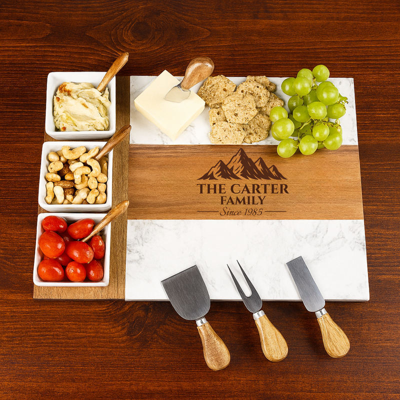 Marble and acacia wood charcuterie board engraved with sample text "THE CARTER FAMILY" and "Since 1985," styled with cheese, crackers, grapes, nuts, and tomatoes in white ceramic bowls, accompanied by three cheese knives with wooden handles.