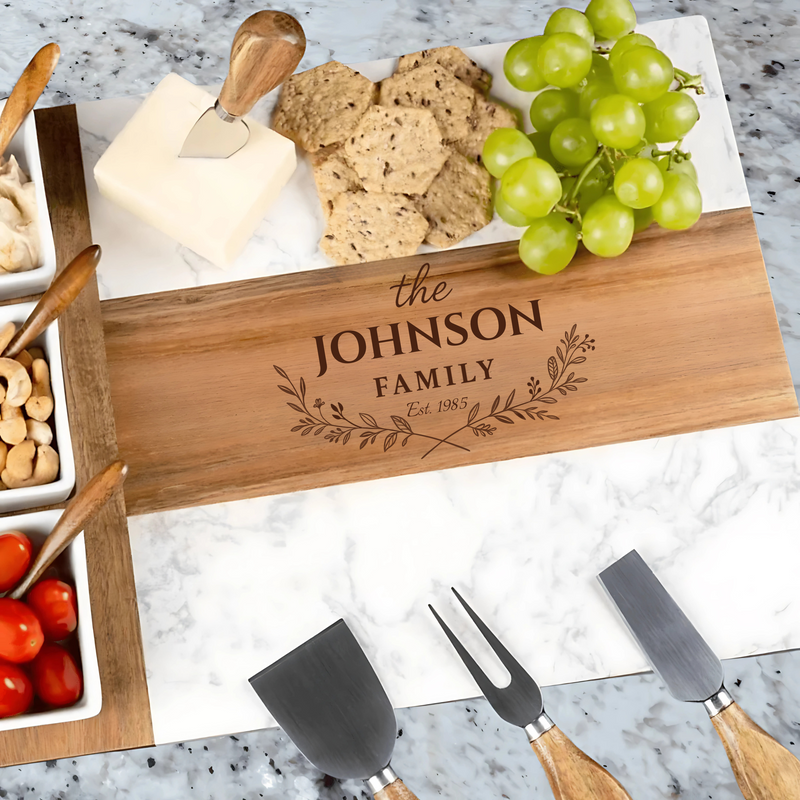 Wooden acacia and marble charcuterie board engraved with sample text "the JOHNSON FAMILY Est. 1985" surrounded by cheese, crackers, grapes, and wooden serving utensils