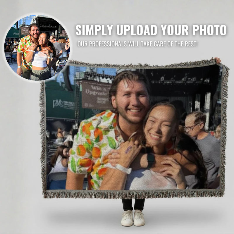 Person holding up a woven photo throw blanket displaying an enlarged image of two people at an outdoor event, with fringe edging visible and a smaller circular photo preview in the upper left corner.