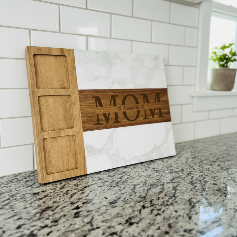 Wooden and marble charcuterie board with laser-engraved sample text "MOM" and "Annabel" arranged in circular pattern, displayed on granite countertop against white subway tile backsplash