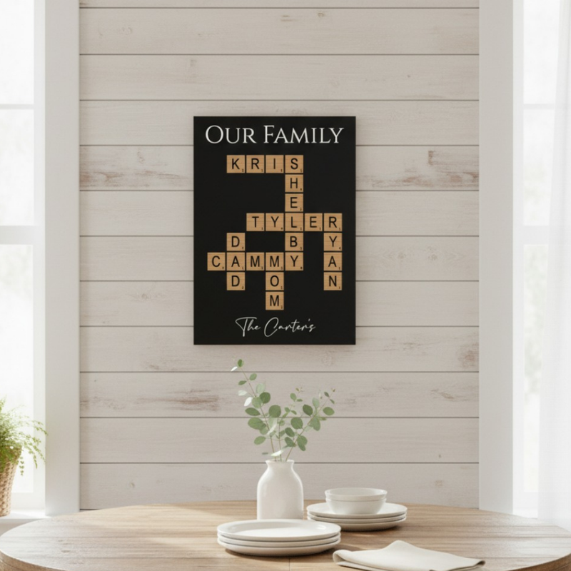 Black wall art with crossword-style layout displaying sample family names "KRIS," "TYLER," "CAMMY," and others, with "The Carters" printed at bottom, mounted on shiplap wall above wooden table with white dishes and eucalyptus.