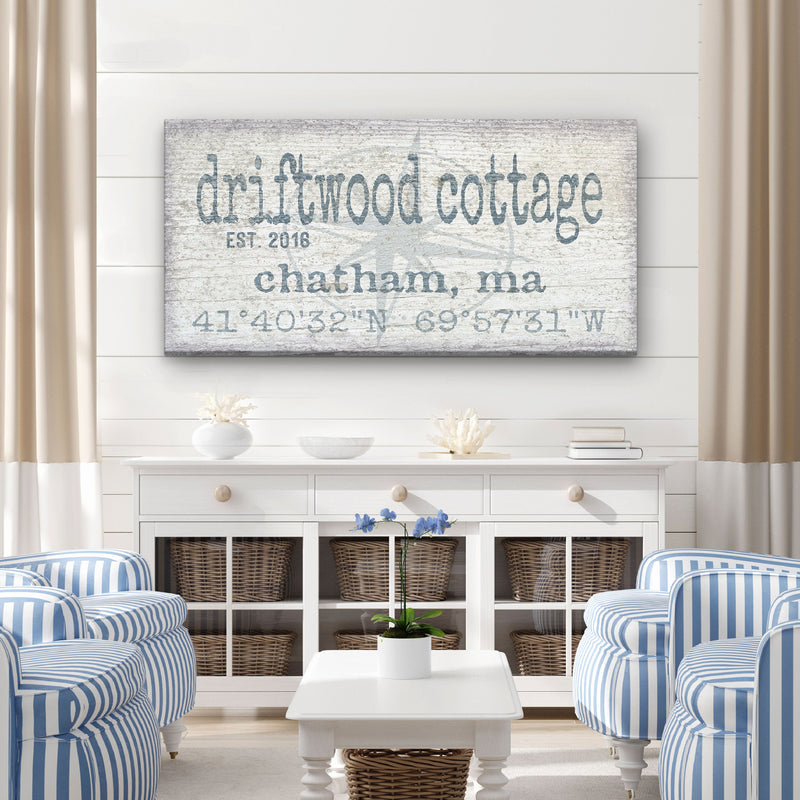 Personalized canvas sign displaying "driftwood cottage" with "EST. 2018," location "chatham, ma," and GPS coordinates, hung above white storage furniture in a coastal-style living room