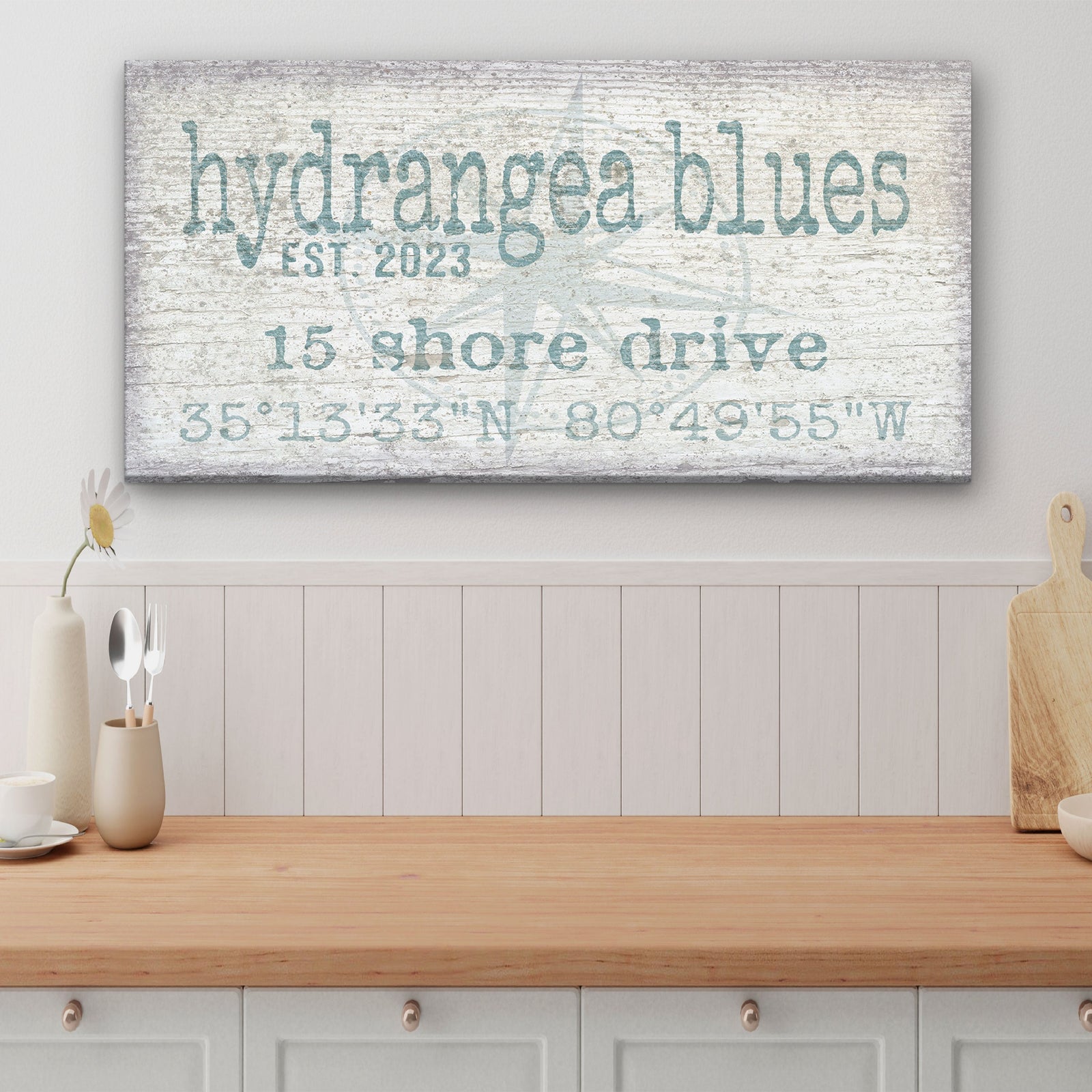 True North Canvas displaying "Rogers" in large bold lowercase above "Family," "Chesapeake, VA," and GPS coordinates with a faded compass rose watermark, in navy blue on a distressed white wood background, mounted above a white credenza in a coastal living room

