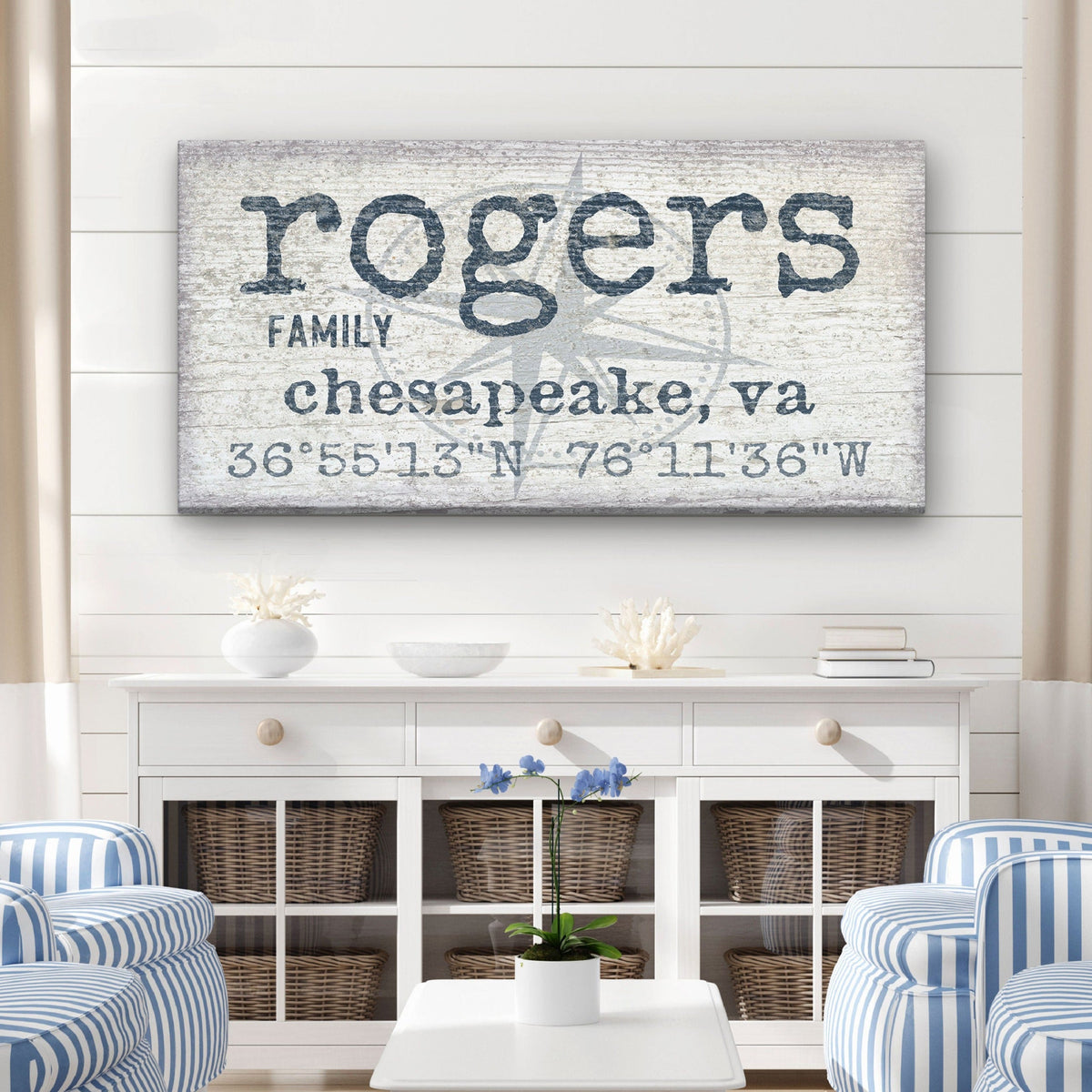 True North Canvas displaying "Rogers" in large bold lowercase above "Family," "Chesapeake, VA," and GPS coordinates with a faded compass rose watermark, in navy blue on a distressed white wood background, mounted above a white credenza in a coastal living room

