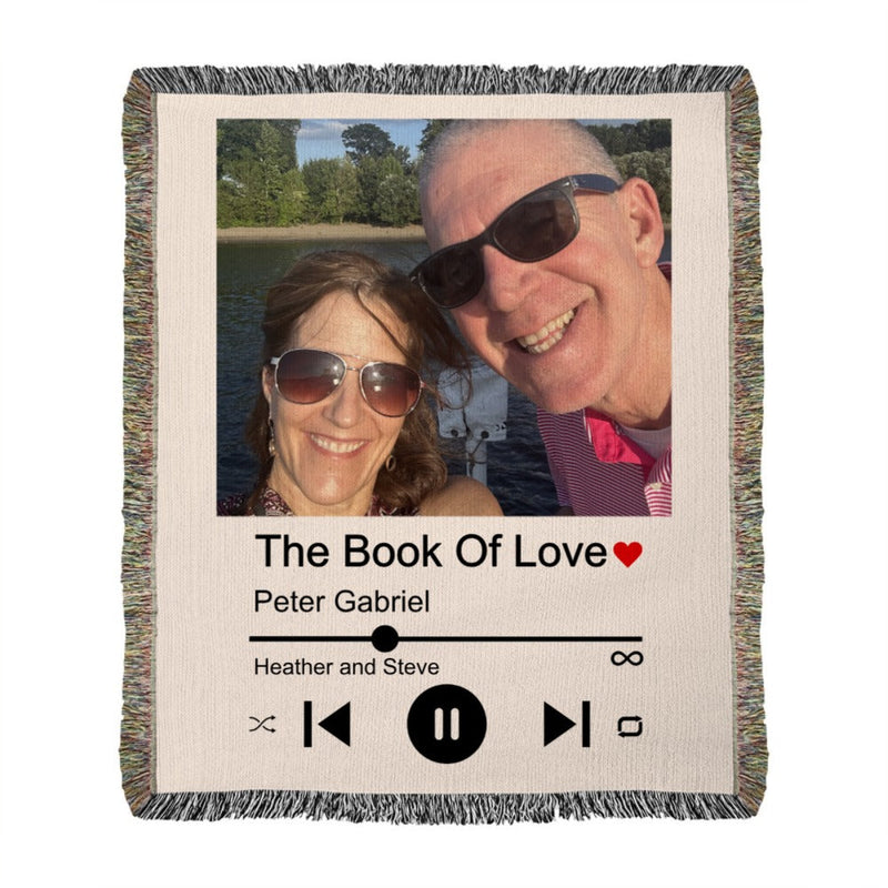 Woven blanket with fringed edges displaying a photo of a couple on a boat, titled "The Book of Love" with a red heart, artist name "Peter Gabriel," couple names "Heather and Steve," and music player controls printed below.