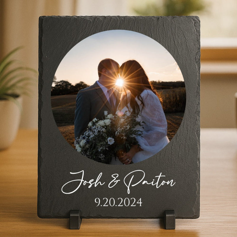 Dark slate picture frame with circular cutout displaying wedding photo of couple kissing at sunset, engraved with sample names "Josh & Paiton" and date "9.20.2024" below the image