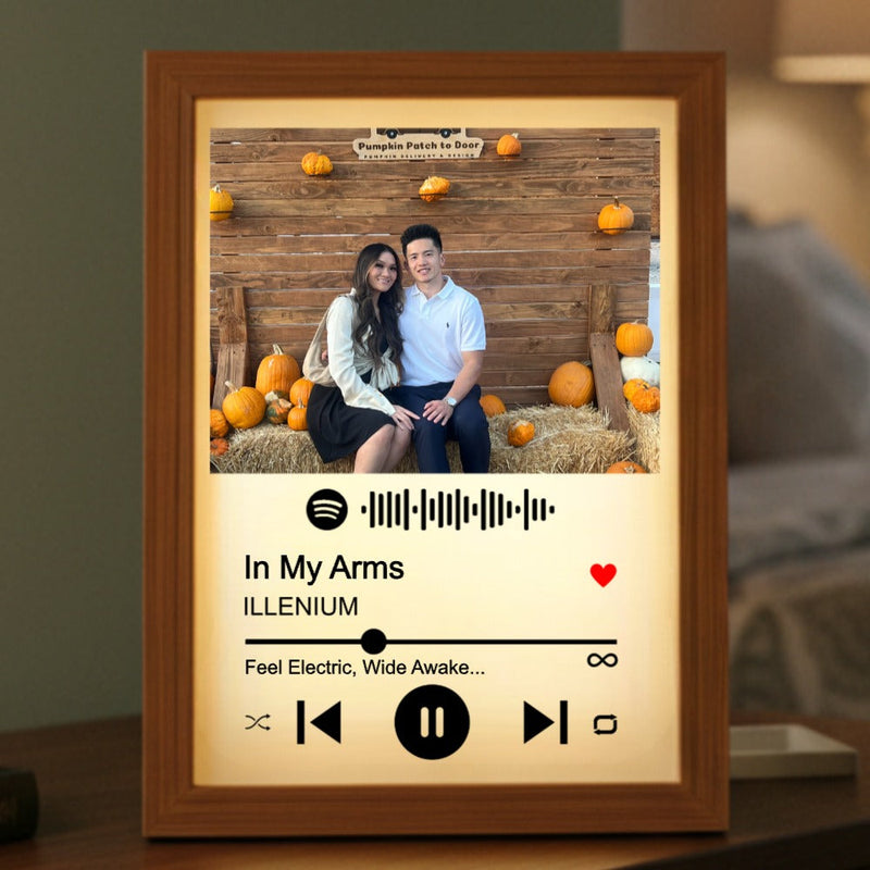 Wood frame with warm backlight displaying a photo of a couple at a pumpkin patch, Spotify code, song title "In My Arms" by ILLENIUM, and music player controls below the image.