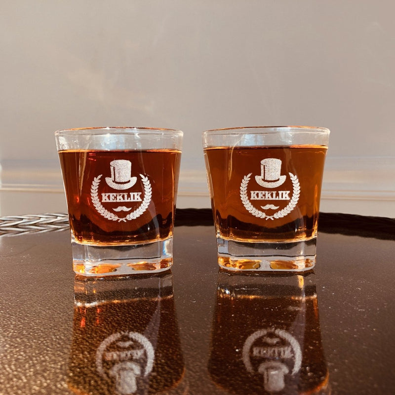 Two rocks glasses filled with whiskey on a dark tray, each engraved with a top hat, laurel wreath, mustache and the name KEKLIK