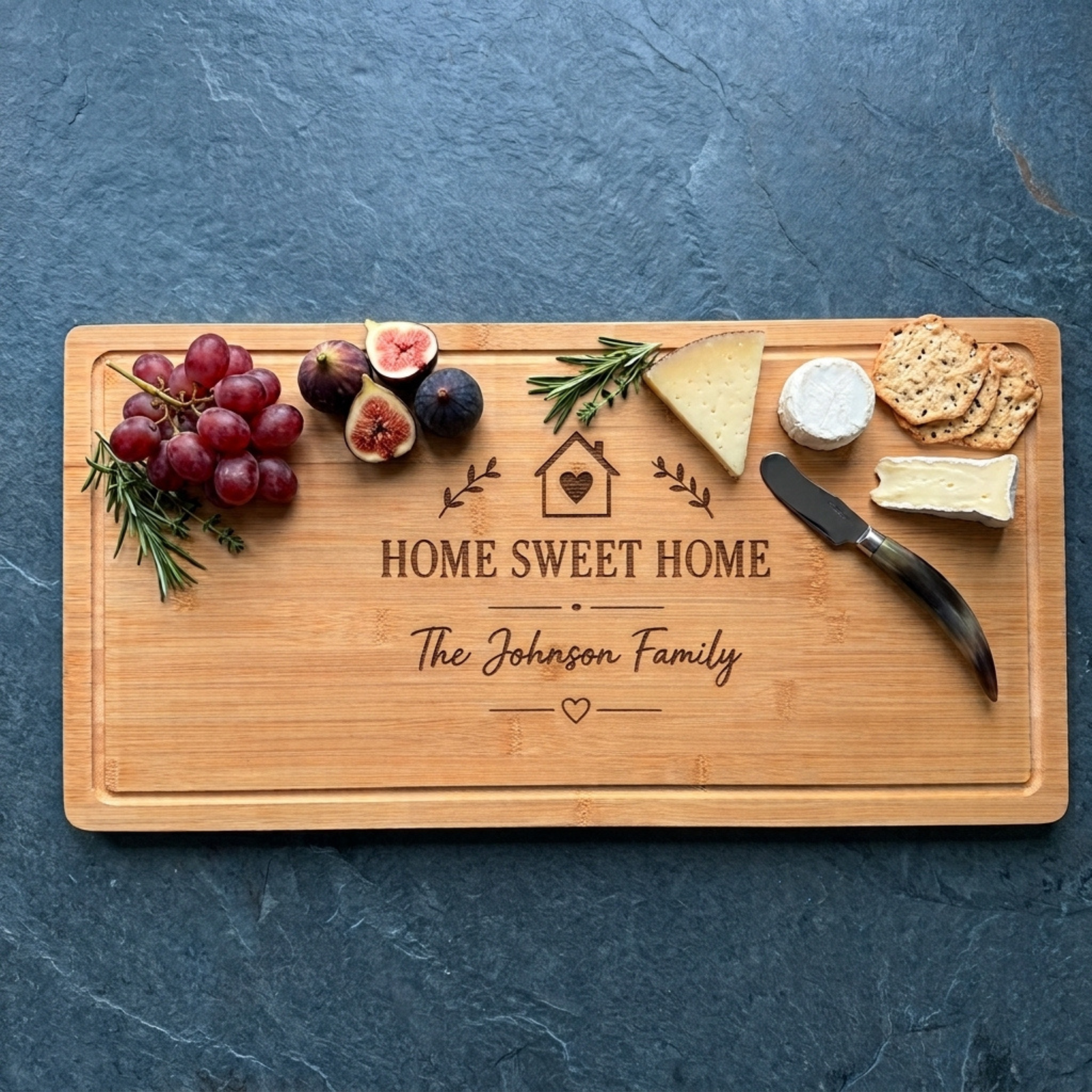 Personalized bamboo housewarming cutting board engraved with a house and heart icon, "Home Sweet Home," and "The Johnson Family," styled with grapes, sliced figs, rosemary, cheese, crackers, and a cheese knife on a dark slate surface. 