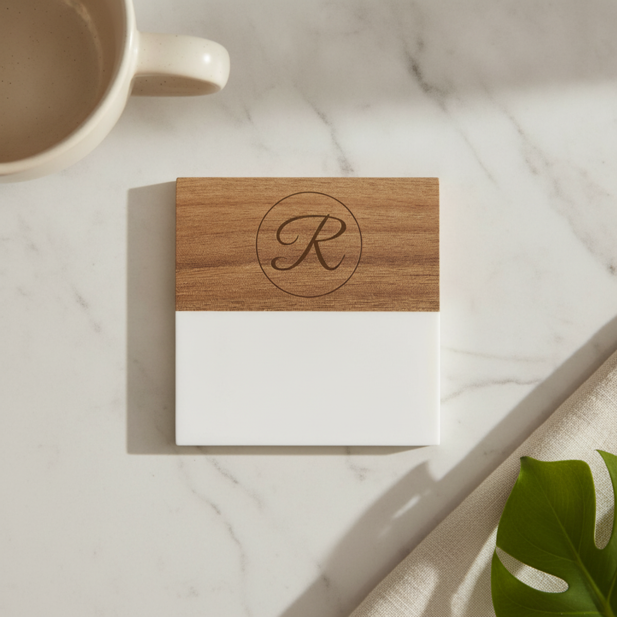 Wooden coaster with sample monogram letter R engraved in circular design, white lower section, on marble surface next to coffee mug