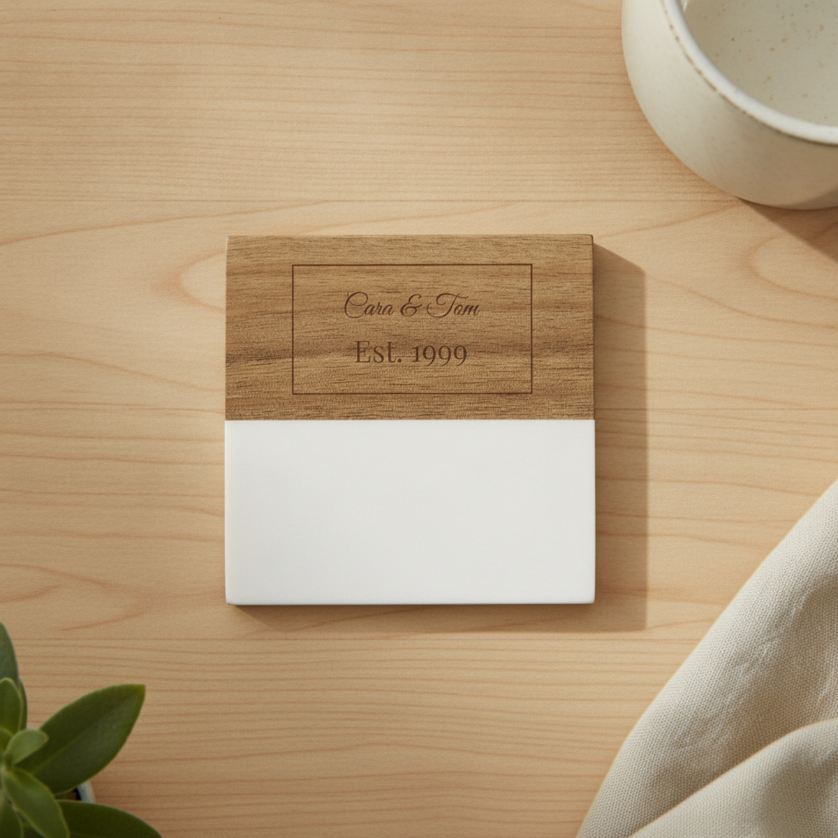 Wooden coaster with engraved sample personalization reading "Cara &amp; Tom Est. 1999" in a rectangular frame, white base section visible below the wood top.