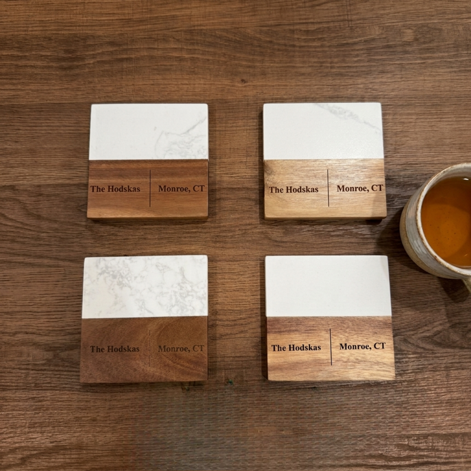 Four personalized marble and wood coasters with sample engraving reading "The Hodskas, Monroe, CT" arranged on wooden surface next to coffee mug