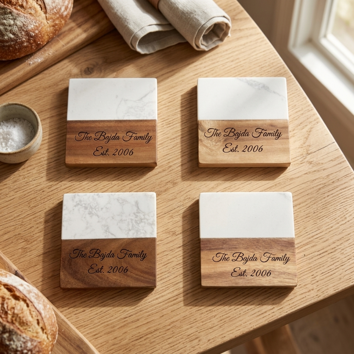 Four square coasters with marble top and wood base engraved with sample family name and establishment year, displayed on wooden table.