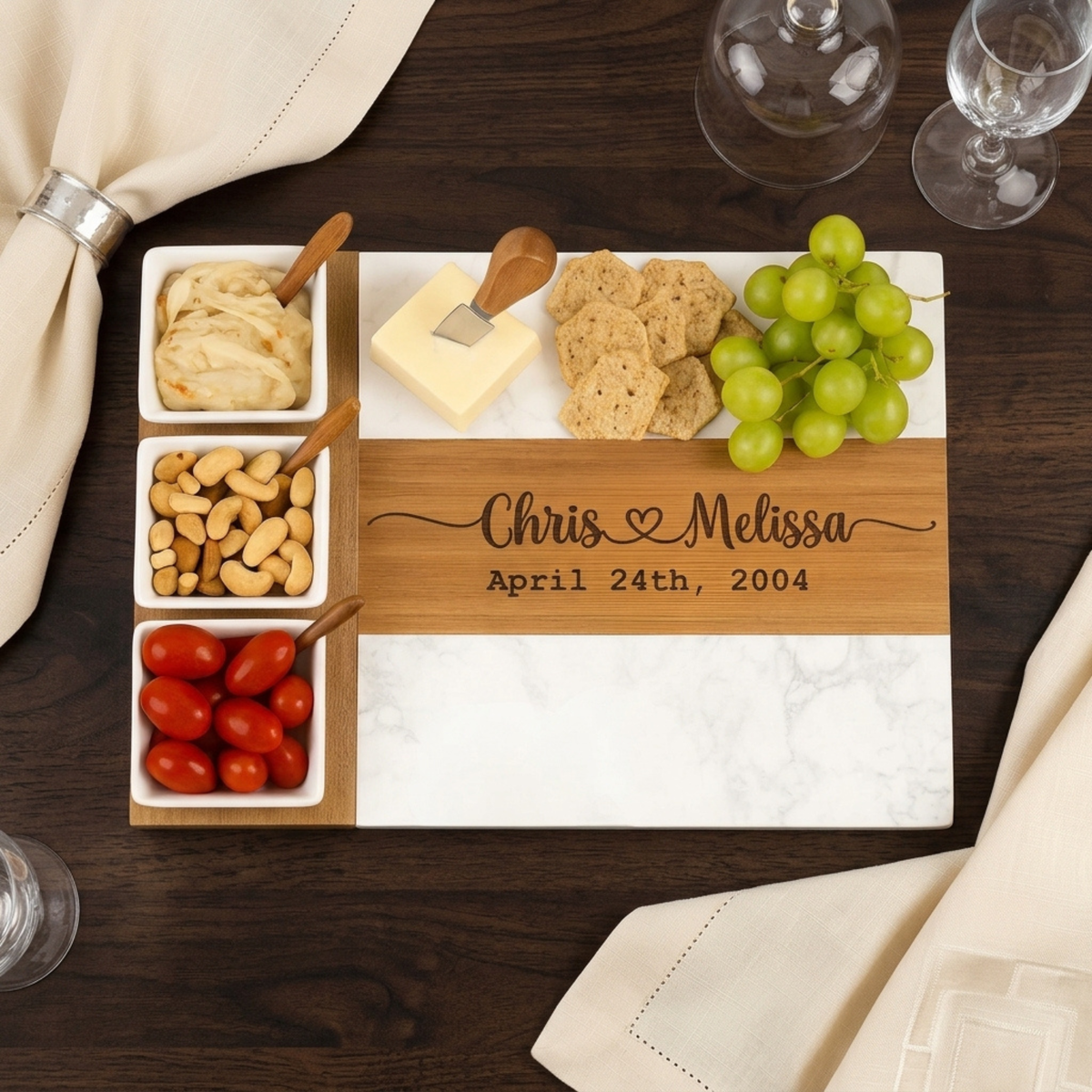 Wooden charcuterie board with engraved sample names and wedding date, featuring white ceramic bowls with nuts, spreads, crackers, cheese, and fresh grapes on dark wood table.