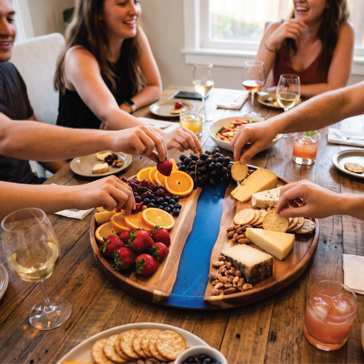 Friends sharing fruit, cheese and crackers served on a round resin river charcuterie board at a dinner table