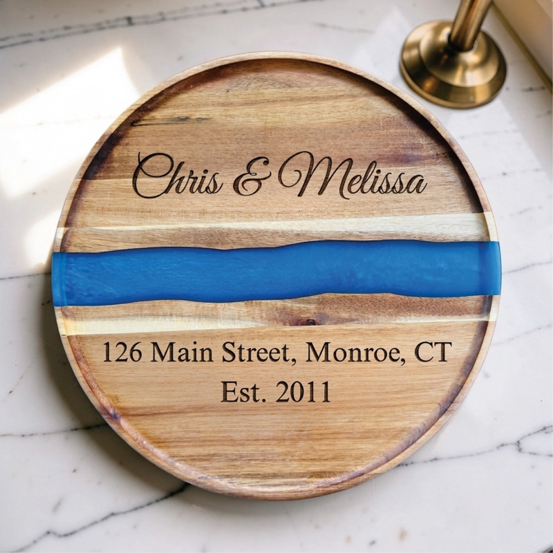 Personalized round acacia charcuterie board with a blue resin river, engraved Chris & Melissa in script with an address and Est. 2011