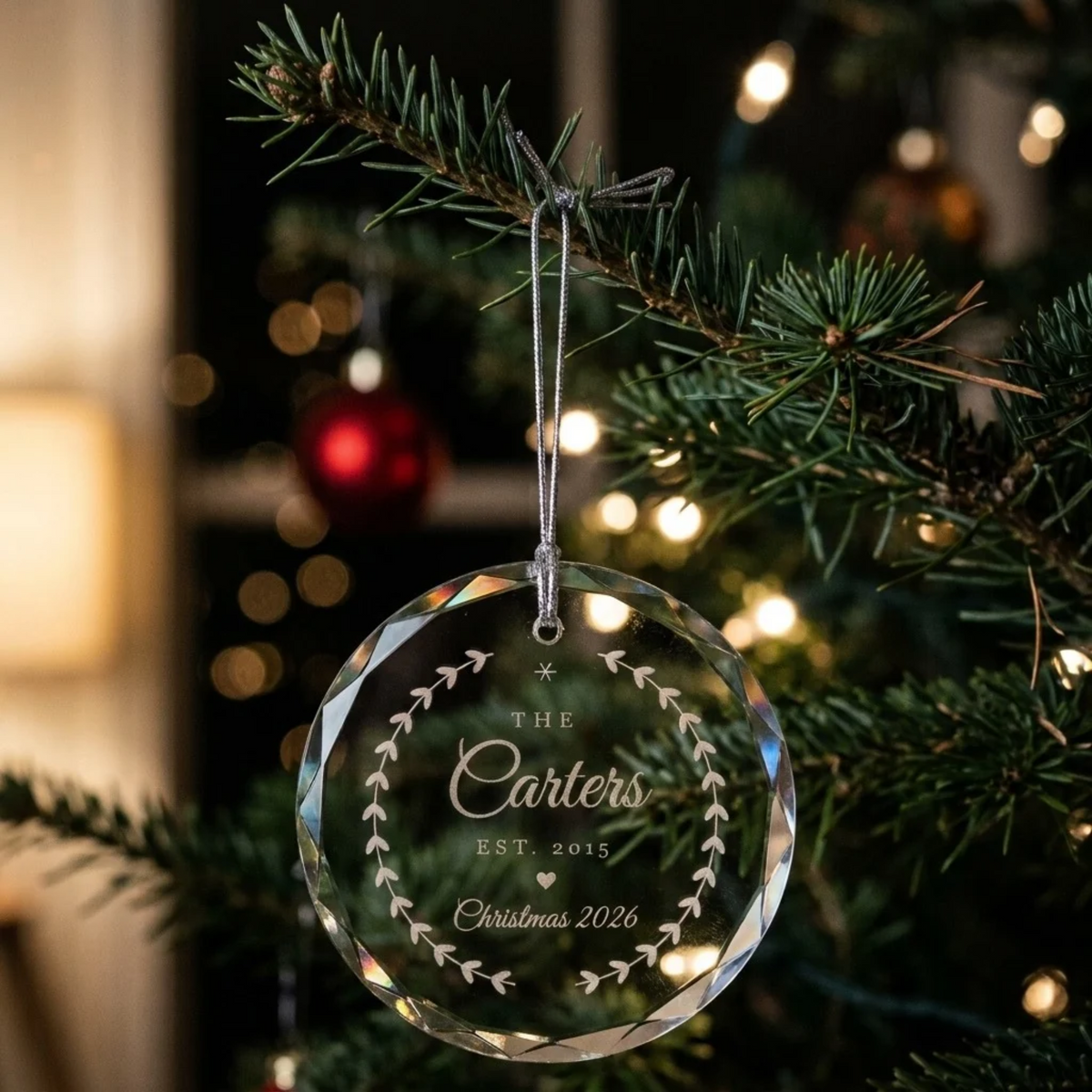 Personalized Family Christmas Ornament in clear faceted glass hanging on a lit Christmas tree, engraved with THE Carters EST. 2015 Christmas 2026 inside a laurel wreath
