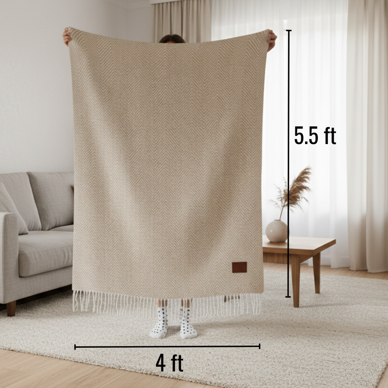 Beige herringbone throw blanket displayed fully outstretched in living room, measuring 4 feet wide by 5.5 feet tall, with fringe trim at bottom and small brown label patch visible