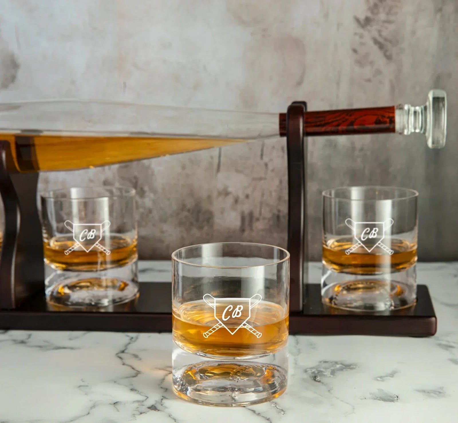 Three whiskey glasses engraved with sample initials "CB" and crossed bats design, filled with amber whiskey, displayed on marble surface with bat-shaped decanter in background
