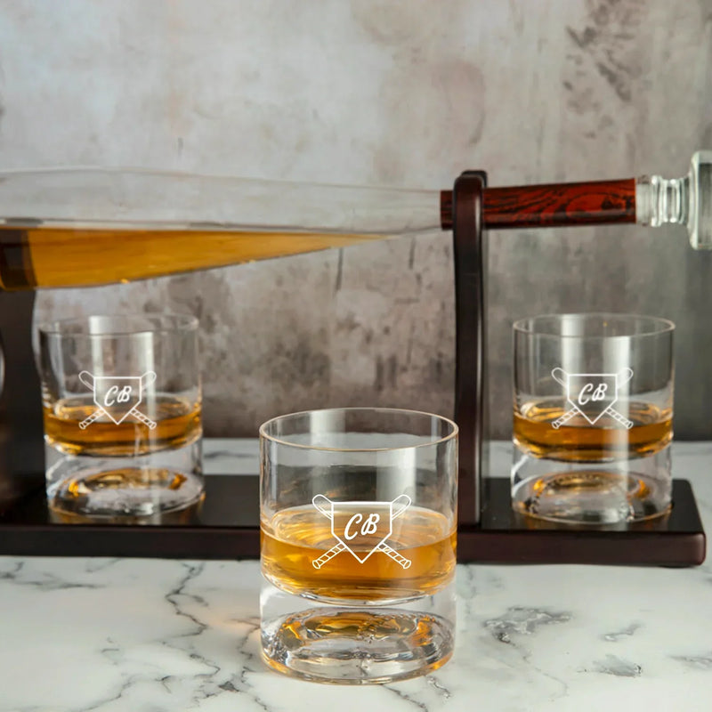 Three whiskey glasses engraved with sample initials "CB" and crossed bats design, filled with amber whiskey, displayed on marble surface with bat-shaped decanter in background