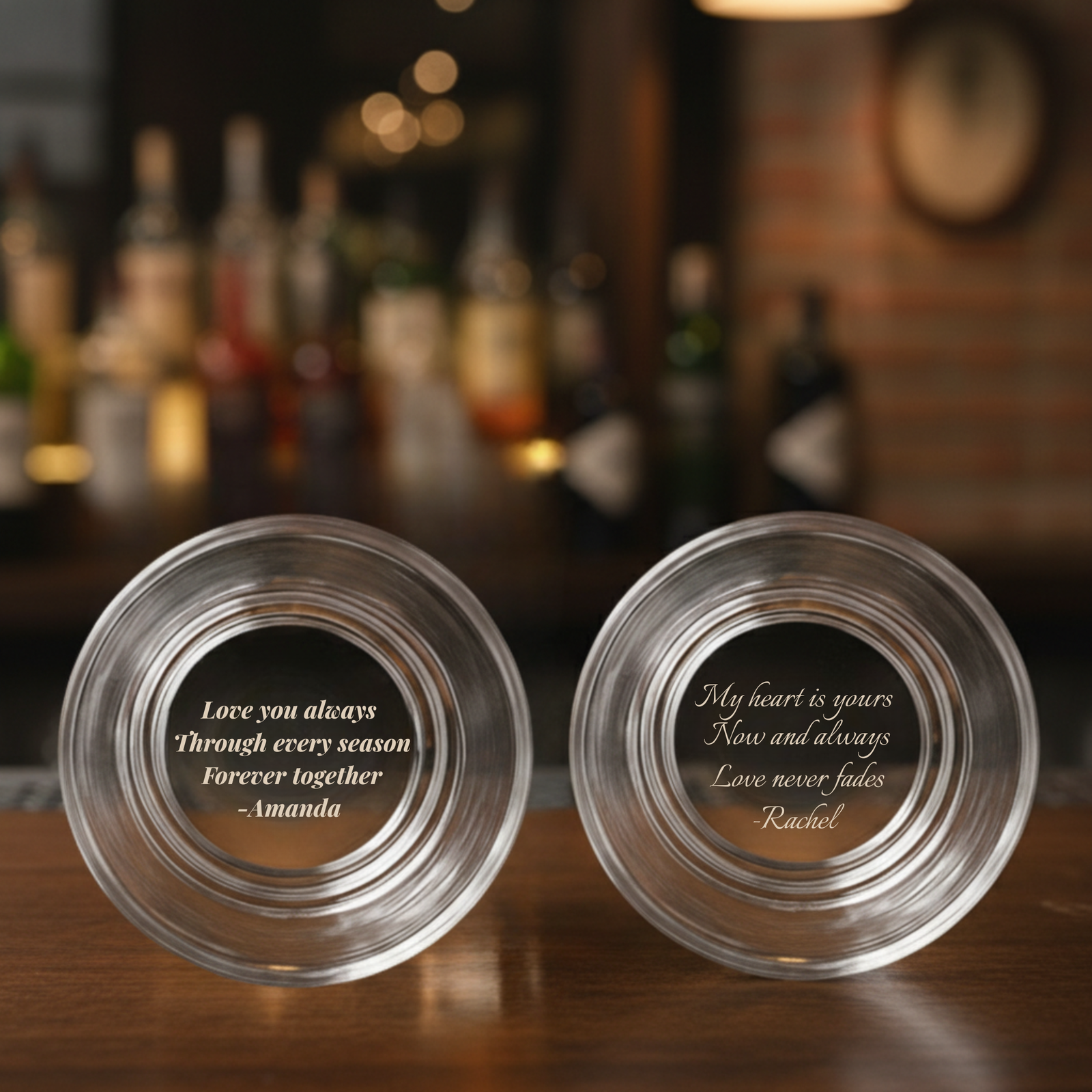 Two personalized whiskey glasses with sample engraved messages at the base, readable when tilted, displayed on a wooden bar counter with bottles blurred in background