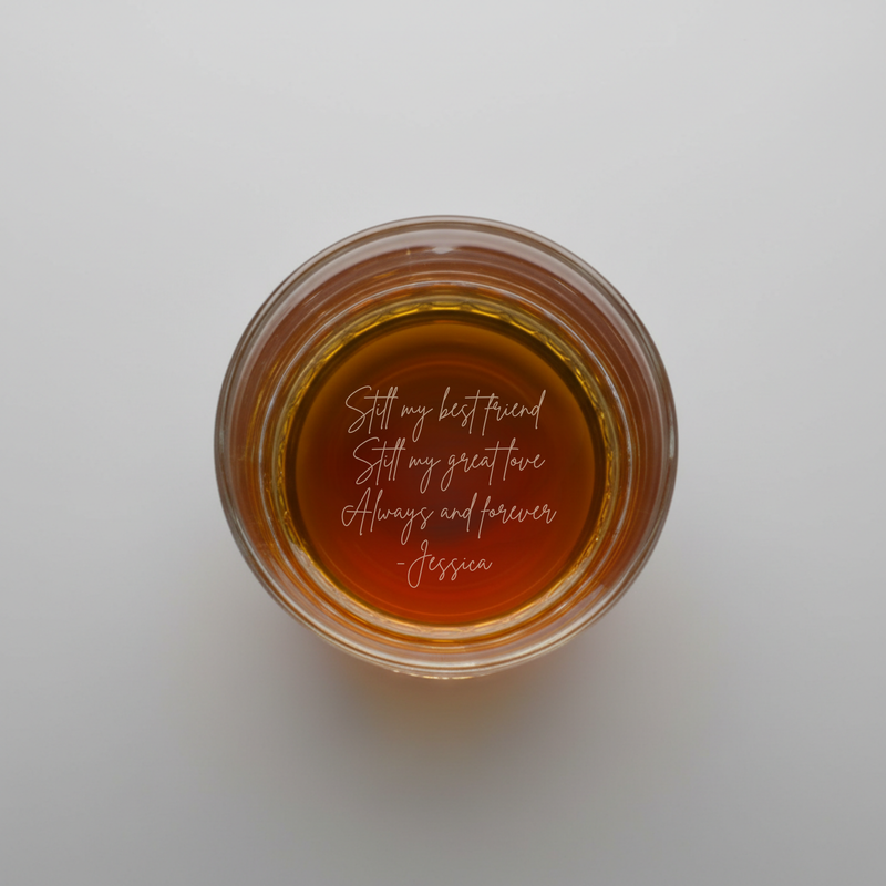 Overhead view of amber whiskey in rocks glass with sample engraved message "Still my best friend / Still my great love / Always and forever / Jessica" visible at bottom