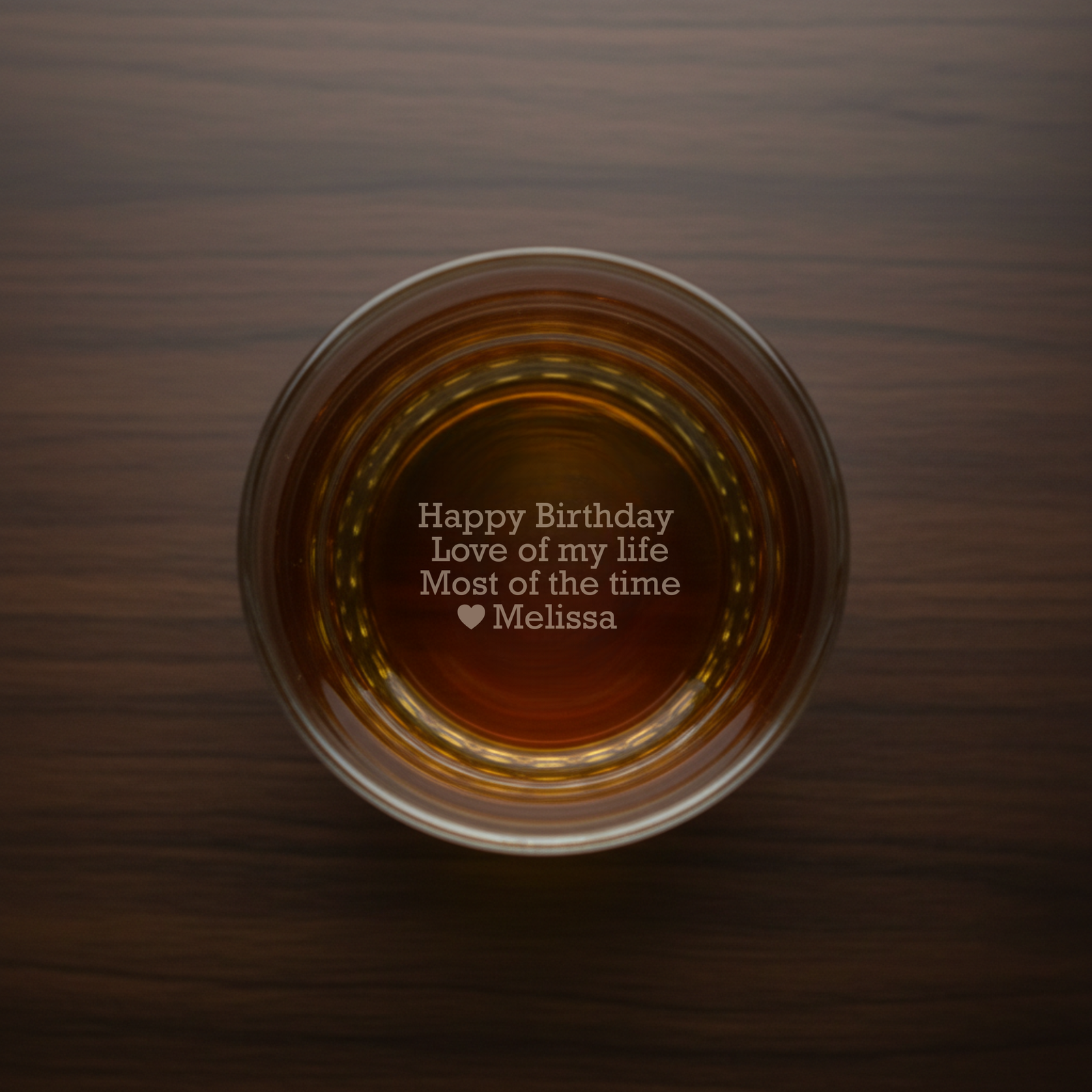 Overhead view of whiskey glass filled with amber liquid, revealing sample engraved message "Happy Birthday Love of my life Most of the time ♥ Melissa" at the base