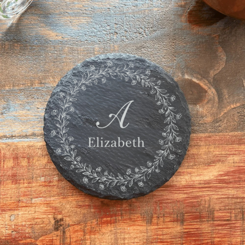 Dark slate coaster engraved with sample initial "A" and name "Elizabeth" surrounded by delicate floral wreath design, displayed on weathered wood surface