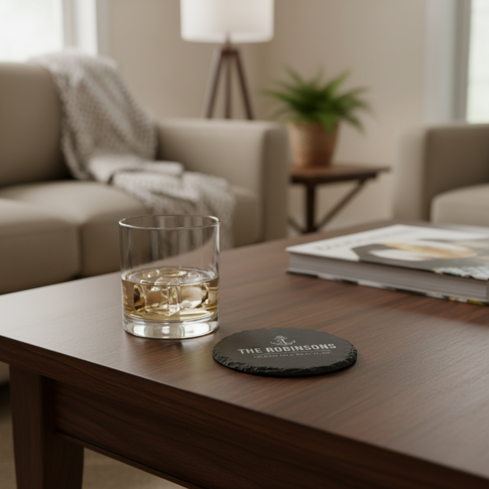 Personalized slate coaster on wooden table with whiskey glass, showing sample engraving "THE ROBINSONS" and location text in living room setting