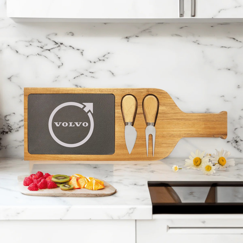 Wooden serving board with slate insert displaying engraved Volvo logo, two wooden cheese spreaders tucked into side slots, styled on marble countertop with fresh fruit and flowers