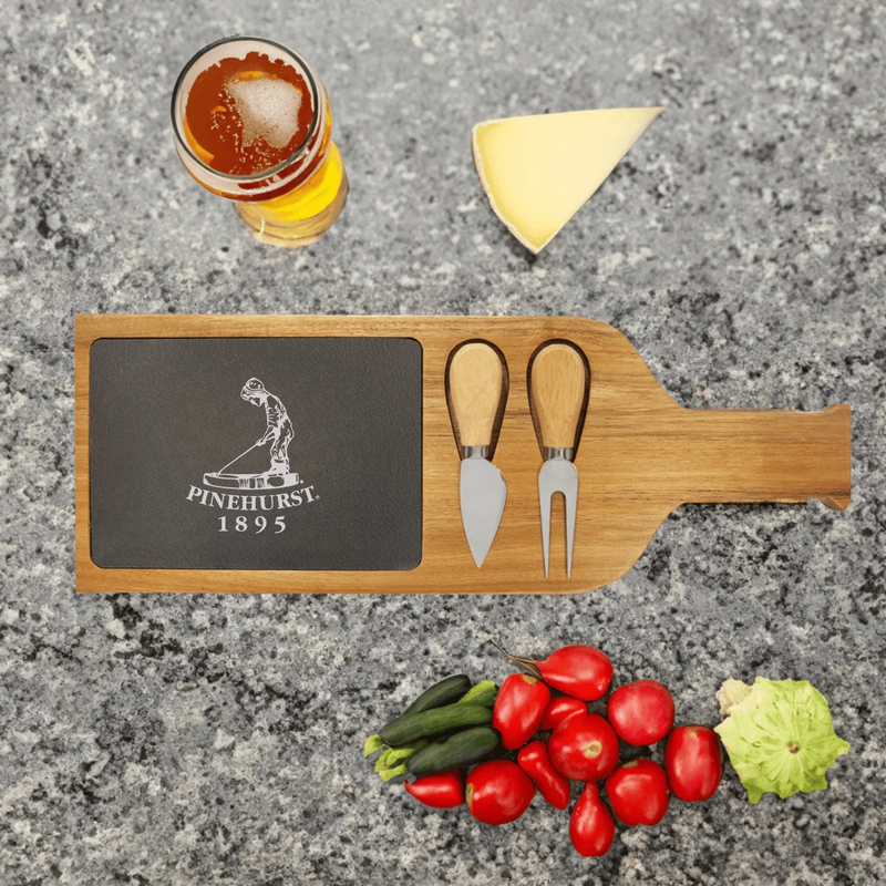 Wooden serving tray with slate insert engraved with sample logo "Pinehurst 1895", two spreader knives, styled with cheese, crackers, and fresh vegetables on concrete surface