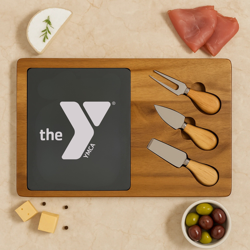 Wooden entertainer's board with slate insert displaying YMCA logo, three cheese spreaders with wooden handles, and surrounding food items including cheese, cured meats, olives, and rosemary sprig
