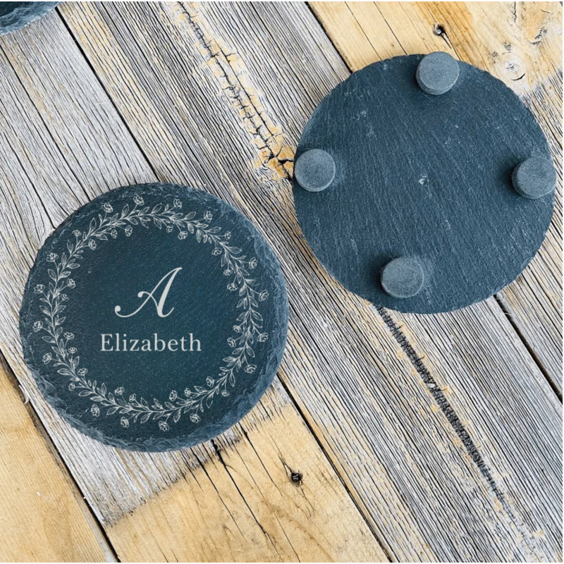 Two slate coasters displayed on weathered wood; left coaster engraved with sample initial "A" and name "Elizabeth" surrounded by delicate floral wreath, right coaster showing plain slate back with gray felt feet.