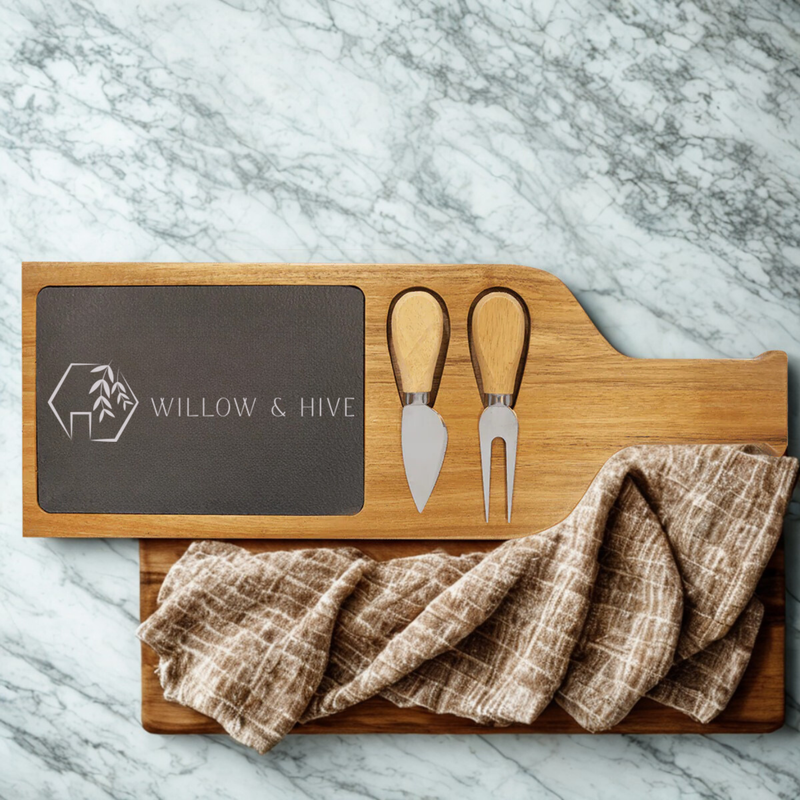 Wooden serving tray with slate insert engraved with Willow & Hive logo, two stainless steel cheese spreaders, and linen napkin draped across base, displayed on marble surface