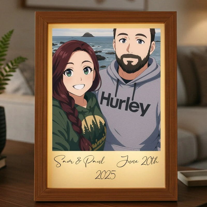 Framed anime portrait light box displaying two illustrated characters with sample names "Sam & Paul" and date "June 20th 2025" printed on cream mat, set on wooden surface with houseplant in background
