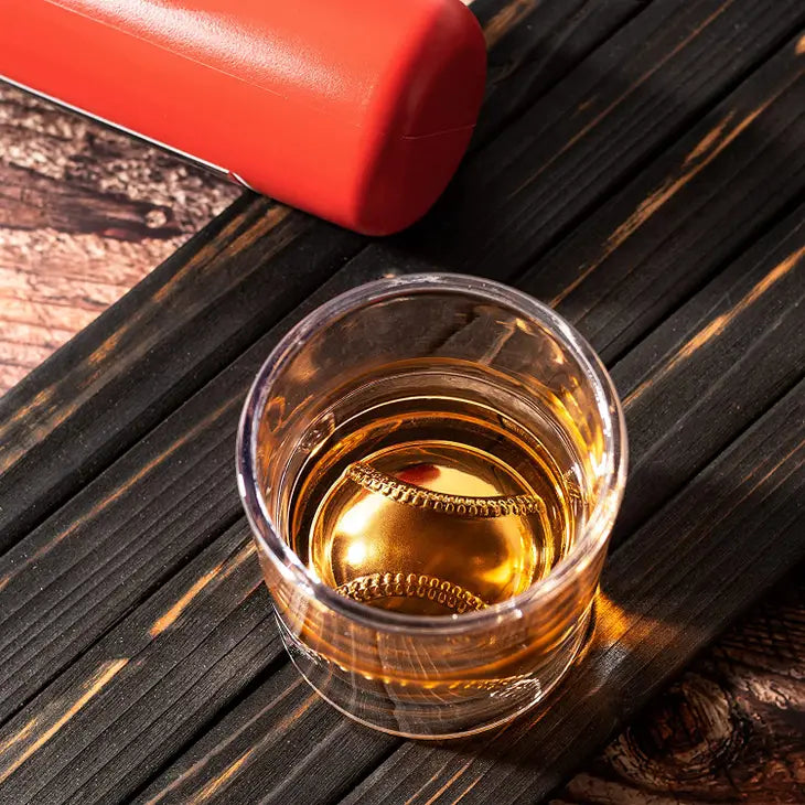 Overhead view of a whiskey glass filled with amber spirit, showing embossed baseball stitching detail on the base, resting on dark wood surface next to red decanter.